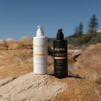 Two bottles of 'dune + kind' products on a rocky surface with a clear blue sky.